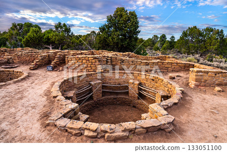 Ruins of Coyote Village in Mesa Verde National Park. A large, circular kiva is prominent in the foreground, with stone walls and trees under a blue sky with clouds 133051100