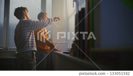 Two Young Male Students Stand Side by Side Next Large, Brightly Light Window With Blinds 133051582