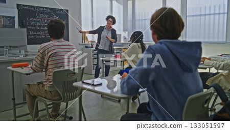 Female Teacher Stands Near Blackboard Explaining Coding Assignment to Group of Students in Bright Classroom Female Teacher Stands Near Blackboard Explaining Coding Assignment to Group of Students in Bright Classroom 133051597