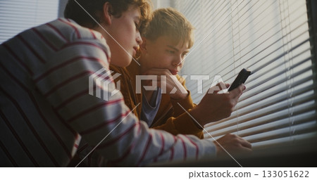 Teen Boys by Bright Classroom Window Scroll Through Phone Together, Exchanging Quiet Comments 133051622