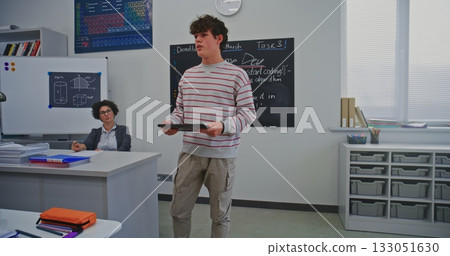 Young Male Student Stands at Front Classroom, Holding Digital Tablet While Delivering Report Young Male Student Stands at Front Classroom, Holding Digital Tablet While Delivering Report 133051630