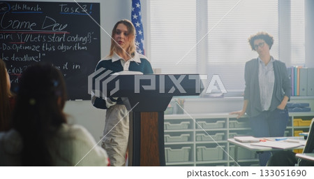 American Female Student Delivers Speech With US Flag in Front of Classmates Sitting Desks 133051690