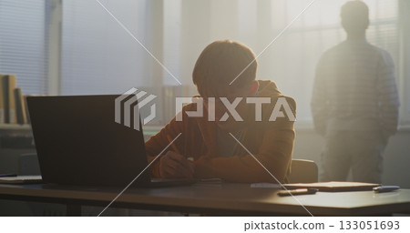 Teenage Boy Sits Desk in Soft Afternoon Light, Focused on Laptop Screen 133051693