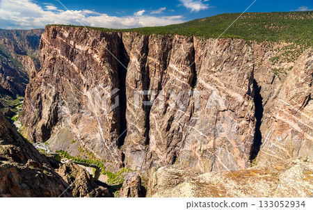 View of the massive Painted Wall in Black Canyon of the Gunnison National Park. White pegmatite dikes create stripes on the dark, sheer cliff. The Gunnison River is visible below 133052934