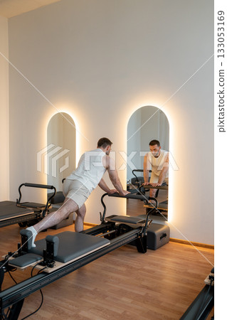 Man training pilates on the reformer bed. Reformer pilates studio machine for fitness workouts in gym. Fit, healthy and strong authentical body. Fitness concept 133053169
