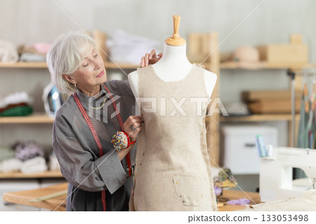 Mature dressmaker working in sewing workshop near mannequin 133053498