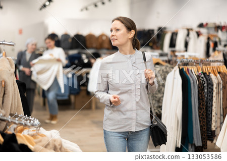 Middle-aged woman watching excitedly large stock of clothes in clothing store Middle-aged woman watching excitedly large stock of clothes in clothing store 133053586