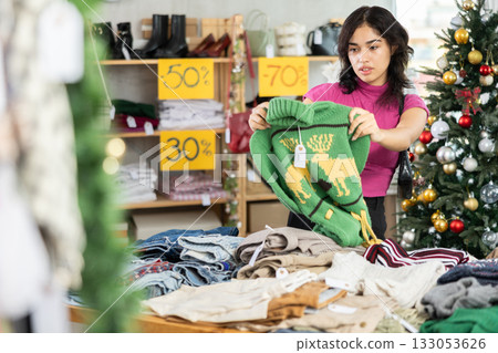 Young Filipino woman choosing sweaters in a store against the background of a Christmas tree 133053626