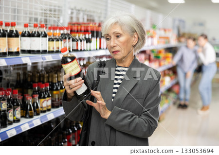 Senior woman customer is thinking about buying spicy and unusual soy sauce. Senior woman customer is thinking about buying spicy and unusual soy sauce. 133053694