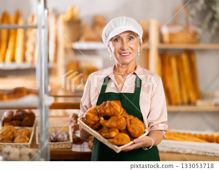 Elderly positive female baker offers hot croissants in uniform in small private bakery Elderly positive female baker offers hot croissants in uniform in small private bakery 133053718