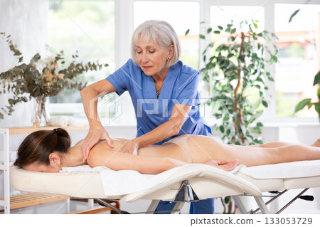 Experienced older professional masseuse doing back massage to young woman Experienced older professional masseuse doing back massage to young woman 133053729