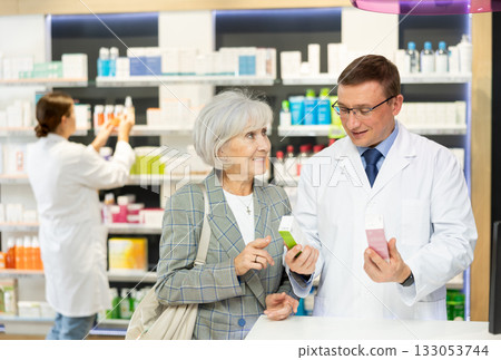 Man apothecary hold package with cream and talks with senior woman client about nuances of care 133053744