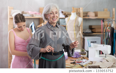 Young tailoress measuring female client in sewing workshop Young tailoress measuring female client in sewing workshop 133053752