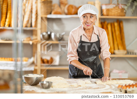 Senior woman works in bakery as baker, cuts dough into portions, form pieces of dough to create buns 133053769