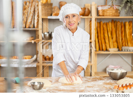 Senior woman works in bakery as baker, kneads dough, works with flour. Senior woman works in bakery as baker, kneads dough, works with flour. 133053805