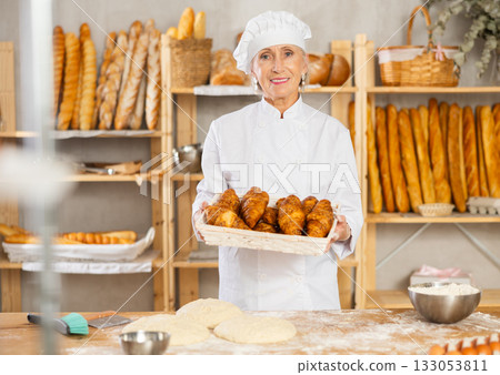 Elderly positive female baker offers hot croissants in uniform in small private bakery 133053811