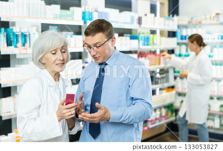Senior woman apothecary hold package with cream and talks with man client about nuances of care 133053827