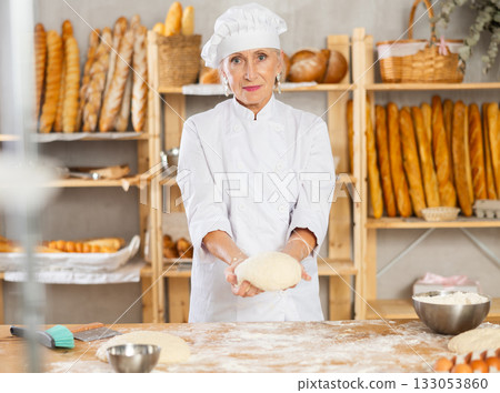 Senior woman works in bakery as baker, kneads dough, works with flour. 133053860