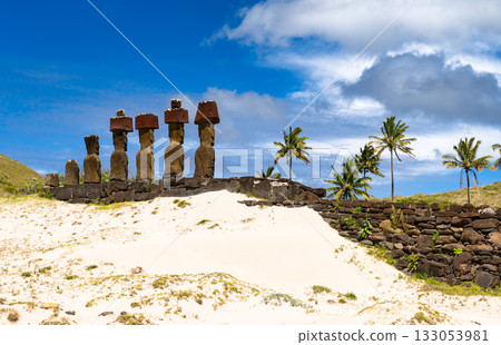 Moai statues with red pukao topknots stand on Ahu Nau Nau at Anakena Beach, Easter Island. White coral sand and palm trees are in the foreground 133053981