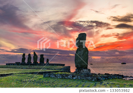 Moai statues at the Ahu Tahai archaeological site on Easter Island (Rapa Nui), Chile. A lone moai stands in the foreground against a dramatic, colorful sunset 133053982