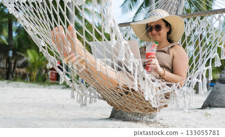 Young woman in a straw hat works on her laptop while lounging in a hammock on a tropical beach enjoying a watermelon shake Young woman in a straw hat works on her laptop while lounging in a hammock on a tropical beach enjoying a watermelon shake 133055781