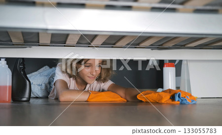 Teen girl in gloves wipes beneath bed in her cozy bedroom with cleaning tools around 133055783