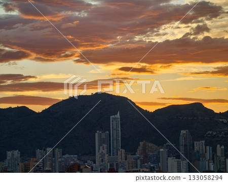 vivid sunset scene, evening sky paints vibrant picture with towering mountains and urban silhouettes 133058294