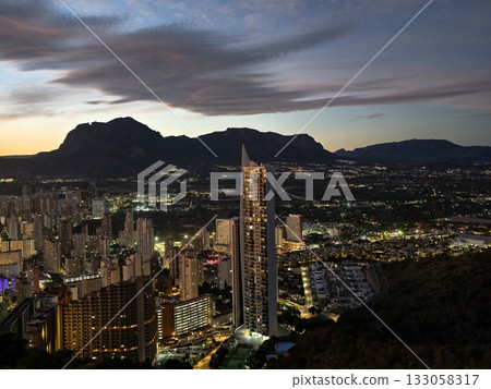 nighttime urban scene with mountains, sunset over city with mountain silhouette and glowing windows nighttime urban scene with mountains, sunset over city with mountain silhouette and glowing windows 133058317