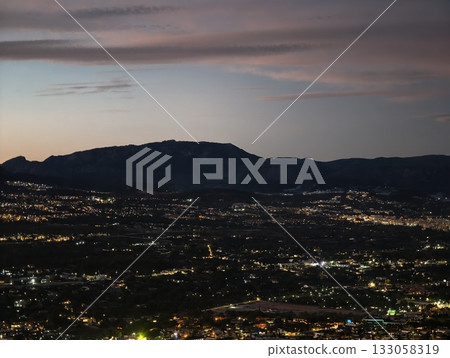 bright cityscape beneath towering mountains, vivid metropolis beyond shadowed mountain ranges 133058319