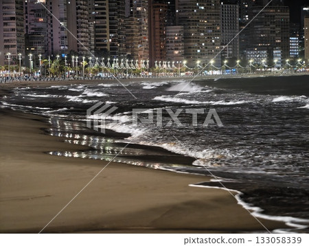 moonlit shore with crashing waves, nighttime seaside view with glowing city lights and rippling surf 133058339