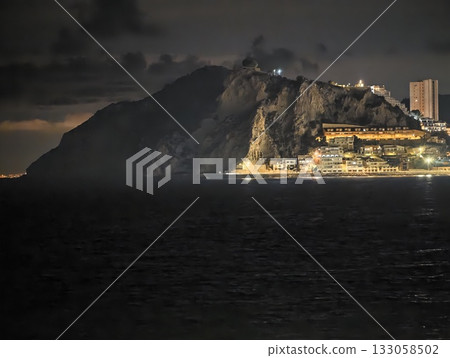 serene nighttime landscape featuring luminous hotel on rugged cliffside with distant city lights 133058502