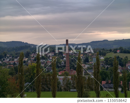 silhouetted trees contrast evening moody sky, hilltop observer watches town and smokestack at dusk 133058642