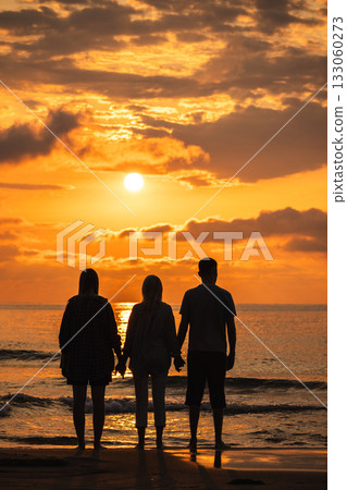 Three people holding hands and watching the sunrise over the sea, standing barefoot on the beach under golden morning clouds. 133060273