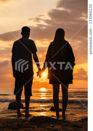 Young couple standing hand in hand on the beach at dawn, enjoying the calm sea and warm golden sunlight reflected on the wet sand 133060278