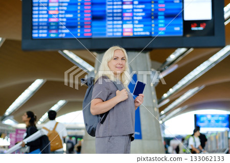 Woman preparing for her flight at an airport terminal while holding a passport and looking towards the departure board Woman preparing for her flight at an airport terminal while holding a passport and looking towards the departure board 133062133