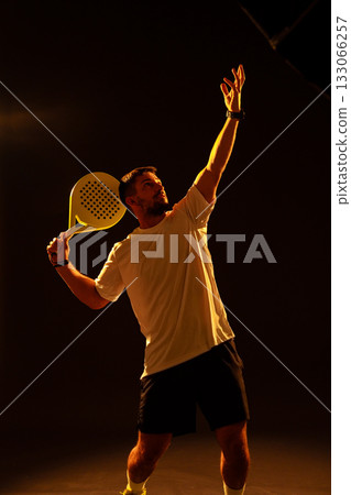 Padel player serves during practice session in indoor court under soft lighting Padel player serves during practice session in indoor court under soft lighting 133066257