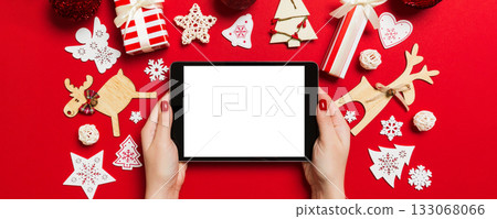 Top view of woman holding tablet in her hands on red background made of Christmas decorations. New Year holiday concept. Mockup 133068066