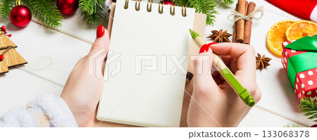 Top view of female hand writing in a notebook on wooden Christmas background. Fir tree and festive decorations. Wish list. New Year concept Top view of female hand writing in a notebook on wooden Christmas background. Fir tree and festive decorations. Wish list. New Year concept 133068378