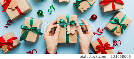 Woman is packing surprise gift for Christmas holidays, sitting at workplace with craft tools, top view. Woman wrapping Christmas gift at colored table 133068606