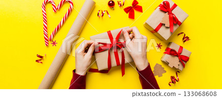 Woman is packing surprise gift for Christmas holidays, sitting at workplace with craft tools, top view. Woman wrapping Christmas gift at colored table 133068608
