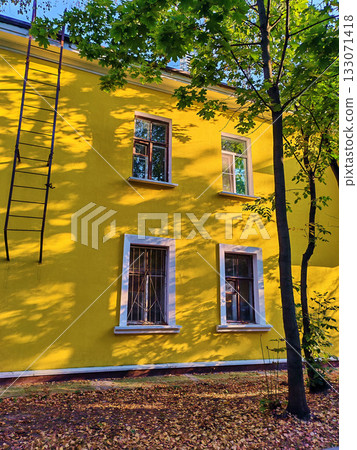 Bright yellow residential building with white window frames and barred lower windows partially shaded by trees. The sunlight filtering through green leaves highlights the fresh renovation, suitable 133071418