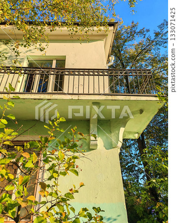 Old green residential building with a balcony featuring a metal railing and decorative supports beneath. The sunlight and greenery emphasize the aged but well-preserved facade, suitable for Old green residential building with a balcony featuring a metal railing and decorative supports beneath. The sunlight and greenery emphasize the aged but well-preserved facade, suitable for 133071425