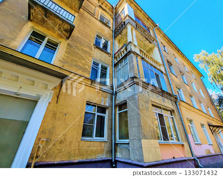 Old yellow residential building with worn balconies and decorative columns showing signs of plaster damage and weathering. The photo highlights facade aging and partial restoration, suitable for Old yellow residential building with worn balconies and decorative columns showing signs of plaster damage and weathering. The photo highlights facade aging and partial restoration, suitable for 133071432