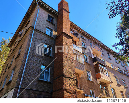 Old multi-story brick building with uneven balconies and a tall red chimney rising along its facade under clear blue sky. The photo conveys the atmosphere of aging Soviet housing, suitable for topics 133071436