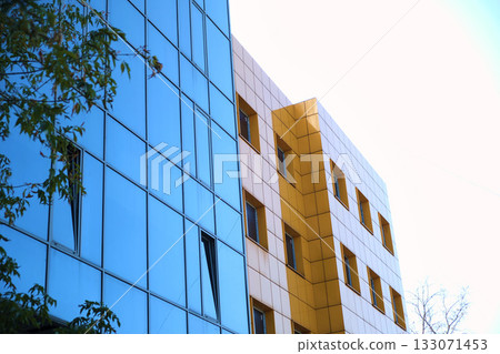 Modern office building with bright yellow facade panels and mirrored glass windows reflecting a nearby high-rise tower. The image illustrates the use of ventilated facades and reflective glazing in 133071453