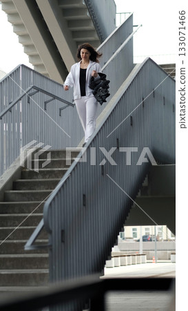 A confident woman in white sportswear walks down an urban staircase, holding Kangoo Jumps shoes. The structured lines of the stairs frame the subject, symbolizing progress and movement. A confident woman in white sportswear walks down an urban staircase, holding Kangoo Jumps shoes. The structured lines of the stairs frame the subject, symbolizing progress and movement. 133071466