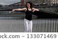 A woman in sportswear gracefully moves near a riverfront, her arms extended in a dynamic motion. The fusion of fitness and urban scenery highlights the energy of an active city lifestyle. 133071469