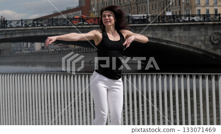 A woman in sportswear gracefully moves near a riverfront, her arms extended in a dynamic motion. The fusion of fitness and urban scenery highlights the energy of an active city lifestyle. A woman in sportswear gracefully moves near a riverfront, her arms extended in a dynamic motion. The fusion of fitness and urban scenery highlights the energy of an active city lifestyle. 133071469