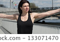 A fit woman warms up outdoors near a city bridge, extending her arms in a power pose. The bright daylight and urban scenery highlight the energy of an active lifestyle. 133071470