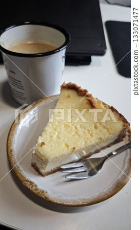 Slice of creamy cheesecake on a plate with a fork next to a cup of coffee. The image conveys comfort, indulgence and a cozy coffee break moment Slice of creamy cheesecake on a plate with a fork next to a cup of coffee. The image conveys comfort, indulgence and a cozy coffee break moment 133071477